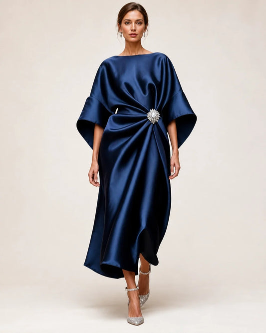 Women's navy satin midi dress with draped waist, crystal flower brooch and wide batwing sleeves, front view.